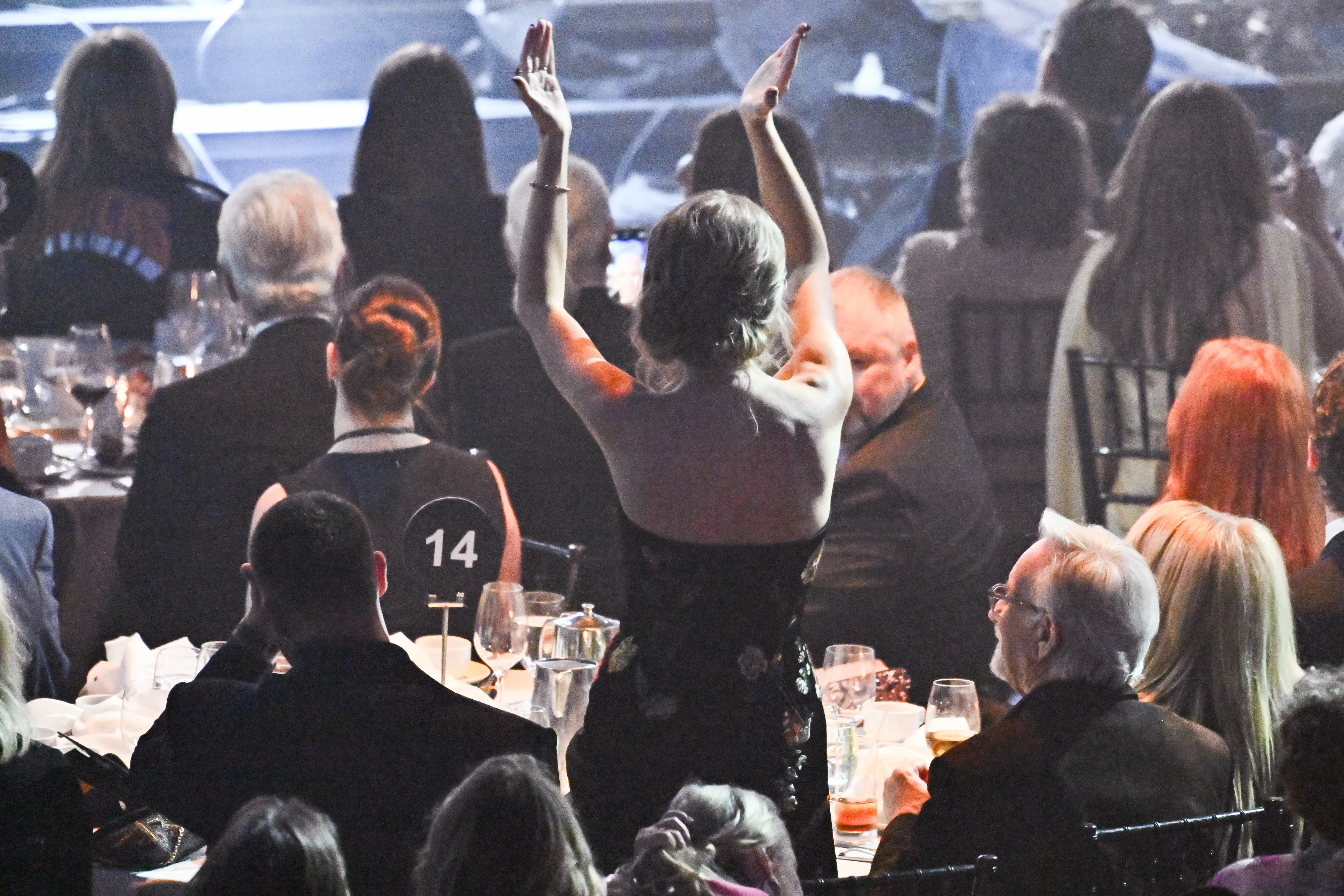 Taylor Swift, viewed from behind, raising her hands in the air amidst a seated audience.