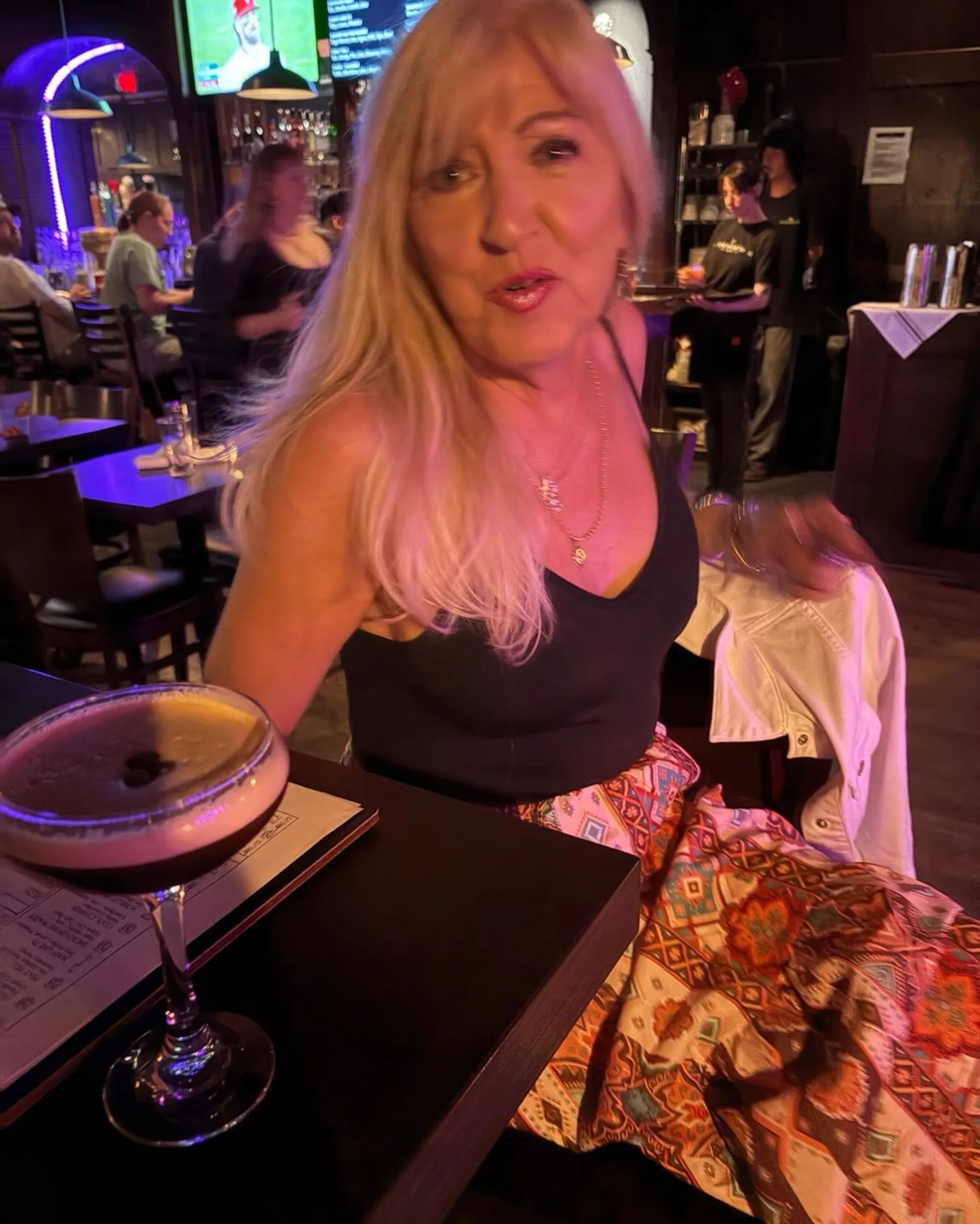 Doree Hepner with long blonde hair and a black top sitting at a table with a drink, in a dimly lit bar.