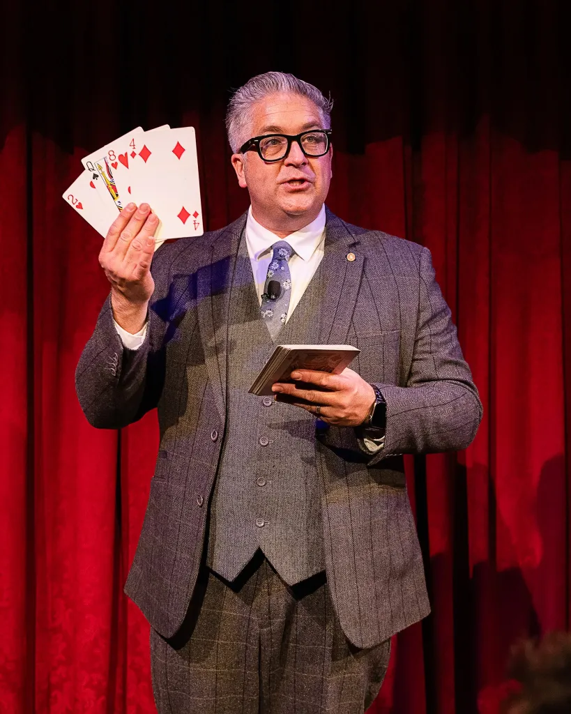 A man in a suit holds up a hand of cards, with a deck of cards in his other hand, in front of red curtains.