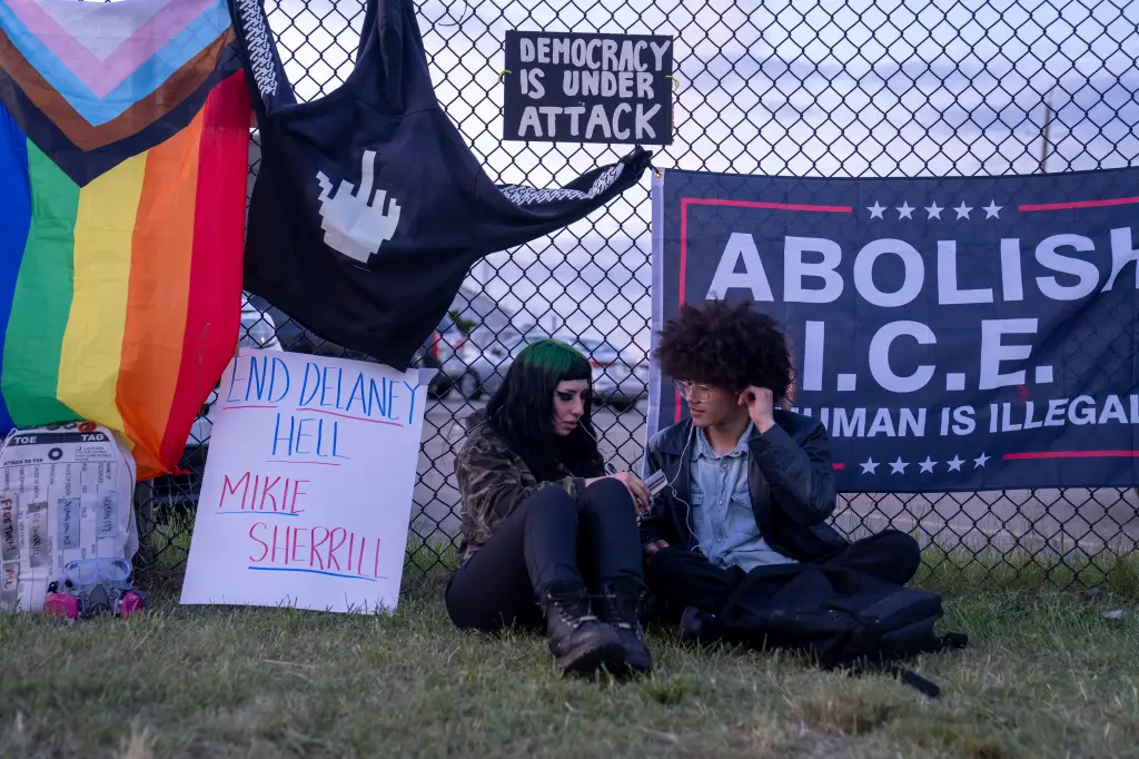 Two people sitting on grass next to a chain-link fence adorned with protest signs, including a Pride flag, a 