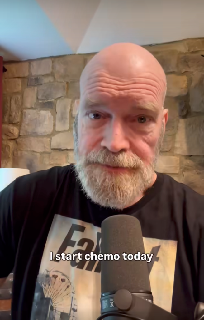 Close-up of Tyler Mane, a bald man with a gray beard, speaking into a microphone, with text overlay 