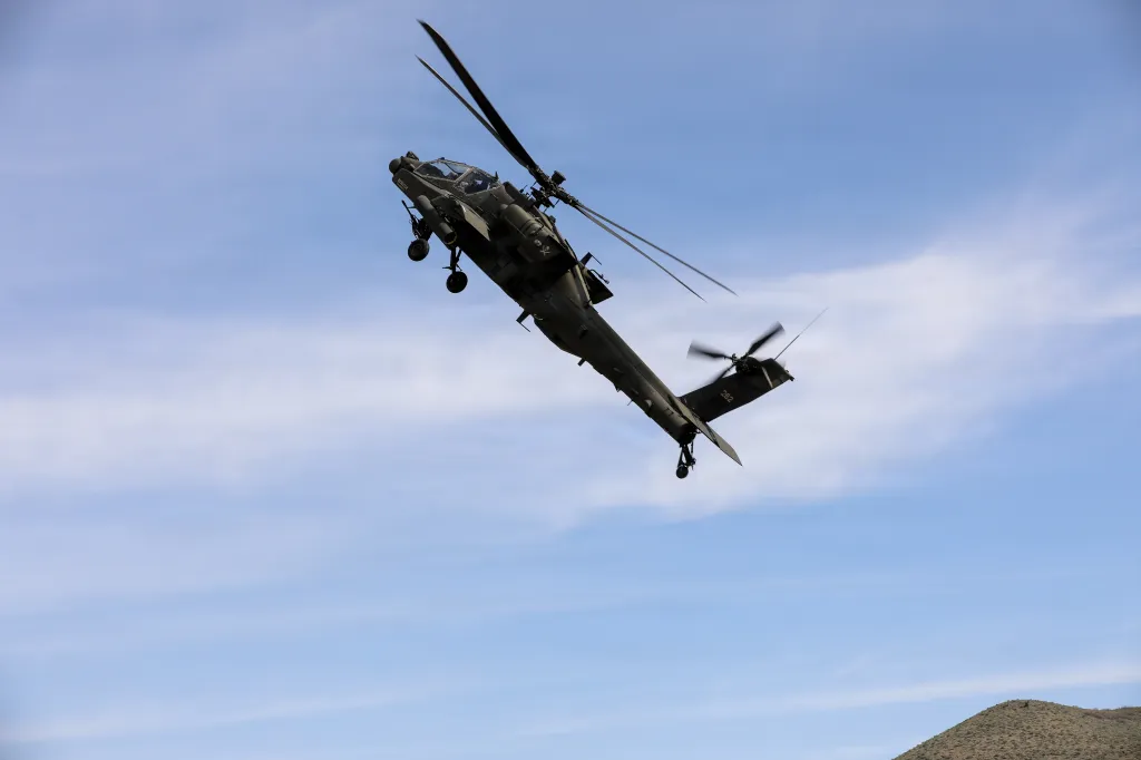 A U.S. Army AH-64 Apache attack helicopter banks sharply during a low-level flight maneuver against a partly cloudy sky.
