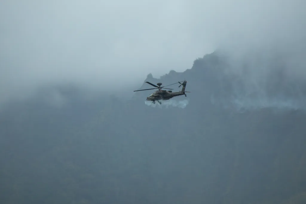 An AH-64 Apache helicopter flying with smoke trailing from its M230 chain gun.