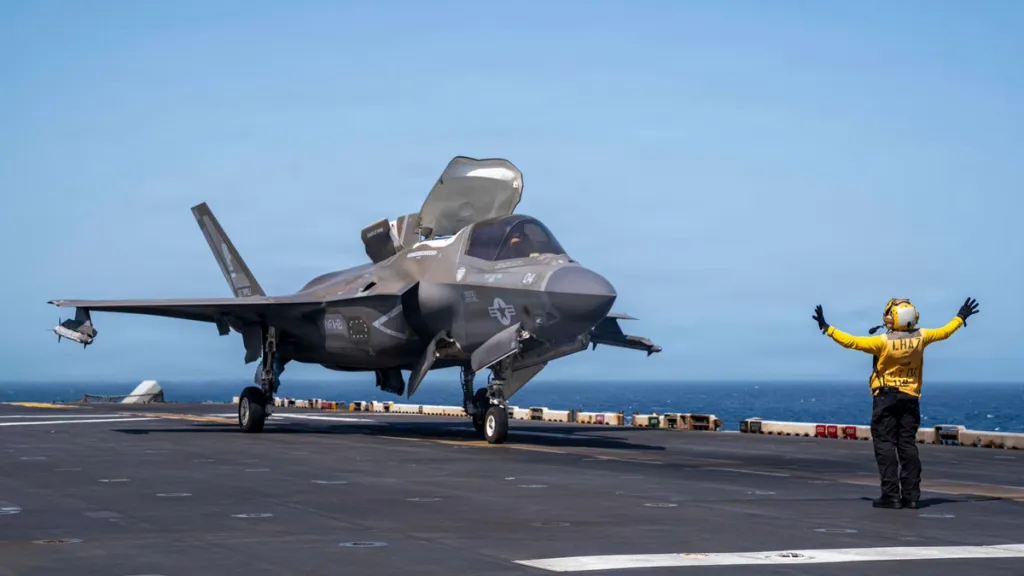 A Marine Corps F-35B stealth fighter jet lands on the USS Tripoli (LHA 7) flight deck with a sailor directing it.