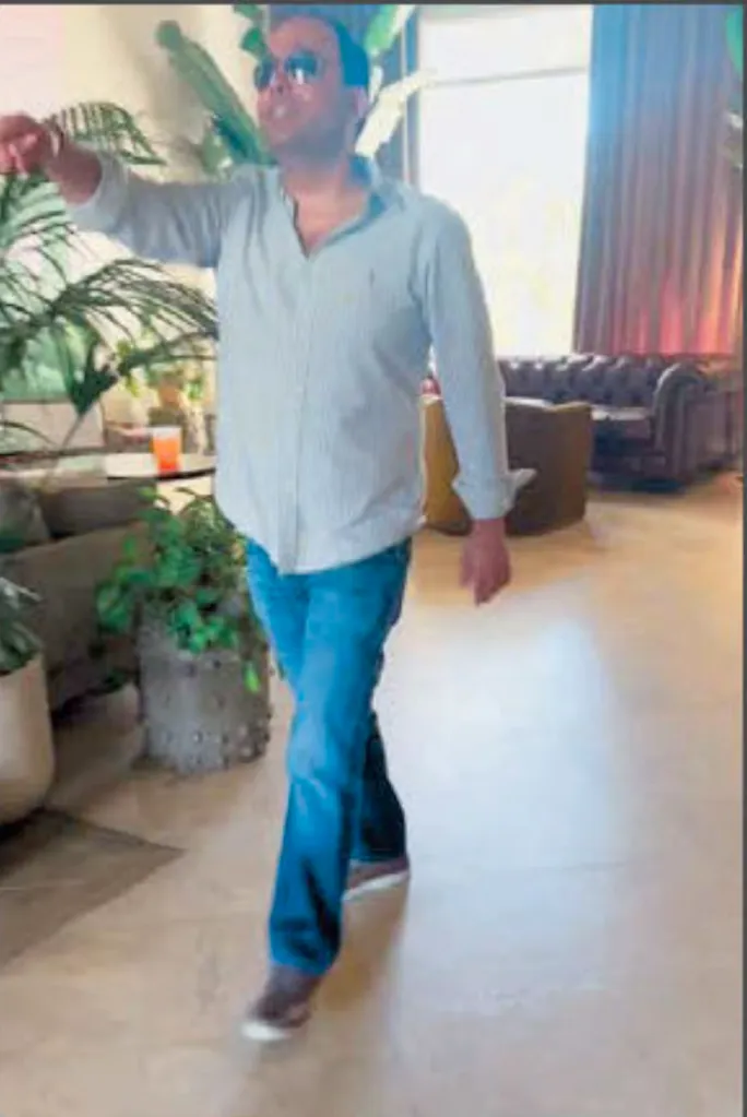 Mahender Makhijani, wearing sunglasses, light shirt, and blue jeans, walking indoors and pointing his left arm forward.