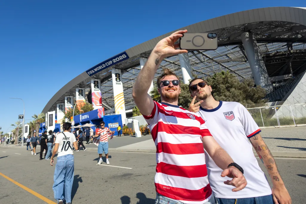 Two male USA soccer fans in jerseys take a selfie outside the FIFA World Cup 2026 stadium.