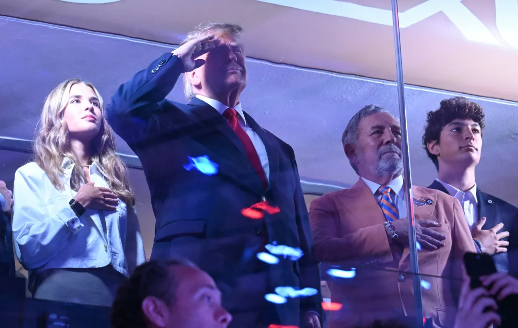 Donald Trump saluting while others stand with hands over their hearts at an NBA game.