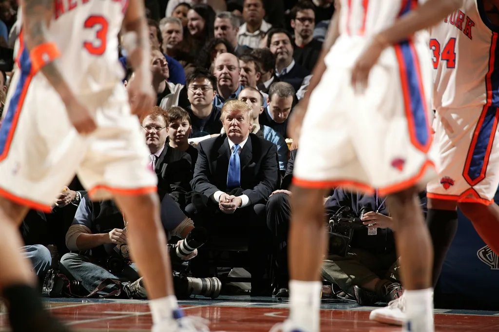 Donald Trump watches a basketball game from the sidelines, with two players in the foreground.