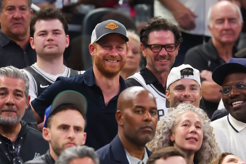 Prince Harry smiling in a baseball cap in the stands at an NBA game.