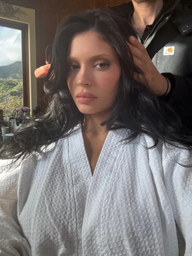 Kylie Jenner wearing a white robe as someone arranges her dark hair.