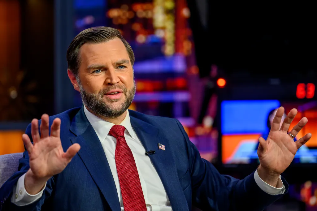 JD Vance speaking on Gutfeld! with hands raised, wearing a blue suit, white shirt, and red tie.