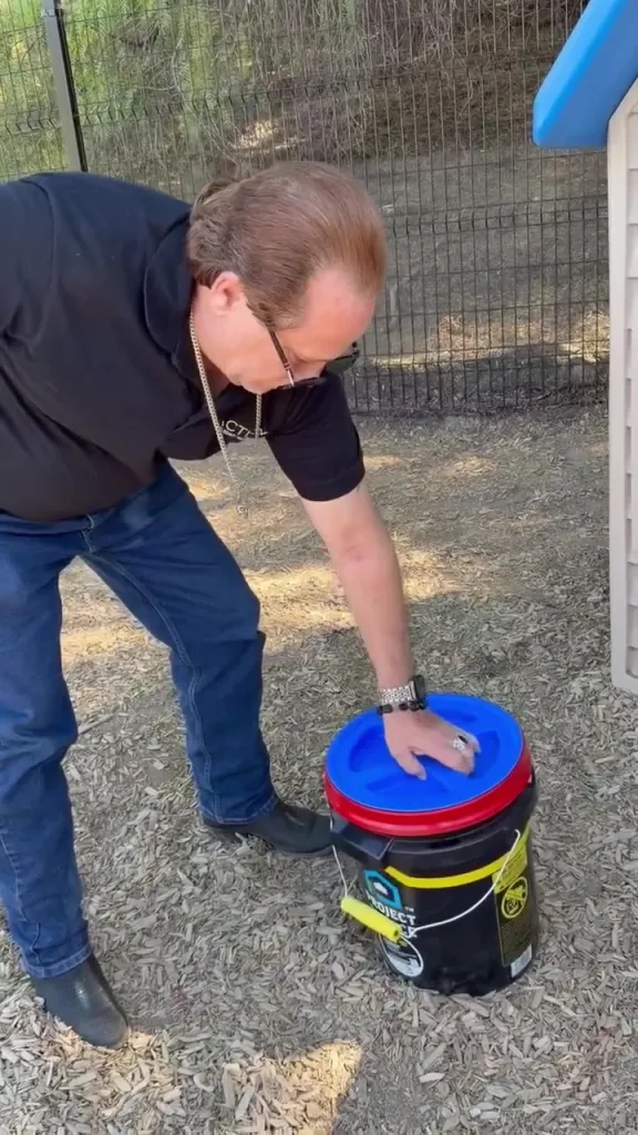 A man in a black shirt and jeans bending over to secure a blue lid on a black bucket.