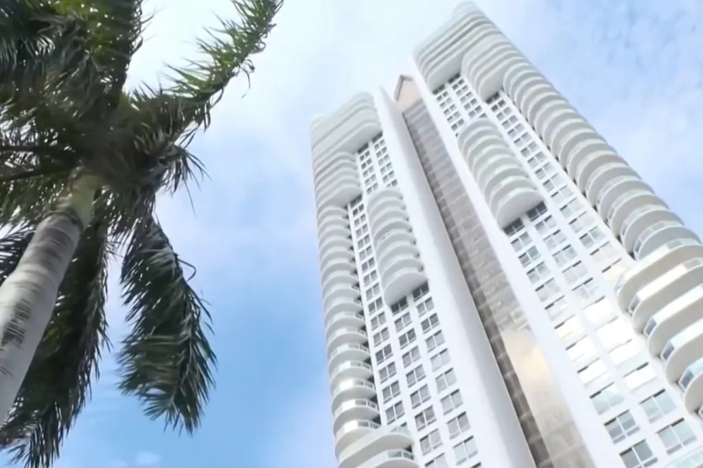 The Akoya Condominium building, a tall white high-rise with many balconies, viewed from below with a palm tree on the left.
