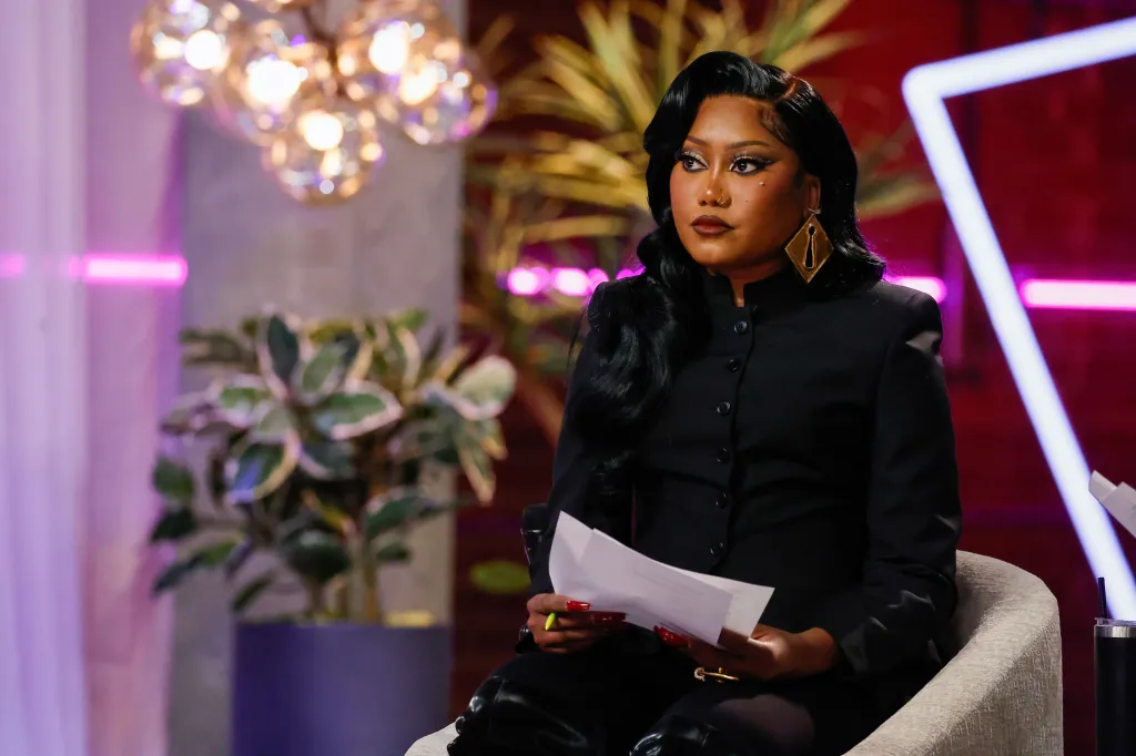 Muni Long sitting and holding papers during the Battles Premiere episode of The Voice.