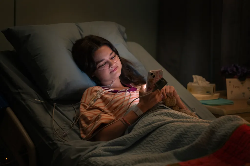 Isabelle in a hospital bed with a nasal cannula and IV, looking at her phone.