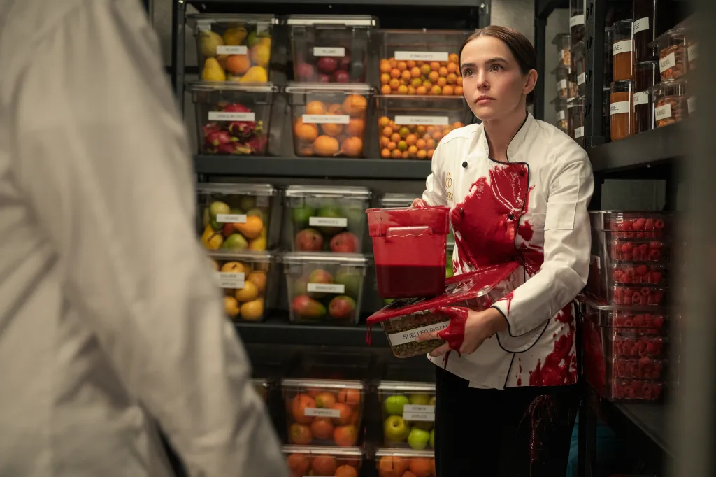 Zoey Deutch as Jill, covered in red liquid, holding a container of red liquid and shelled pistachios, while standing in front of shelves of various fruits and nuts in storage.