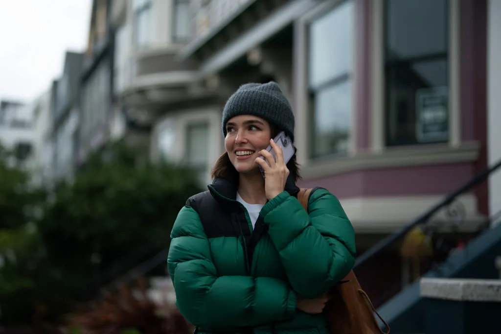 Zoey Deutch as Jill on the phone, smiling, wearing a green puffer jacket and a gray beanie.