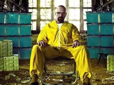 Walter White in a yellow hazmat suit sitting in a room with stacks of cash and blue crystals.