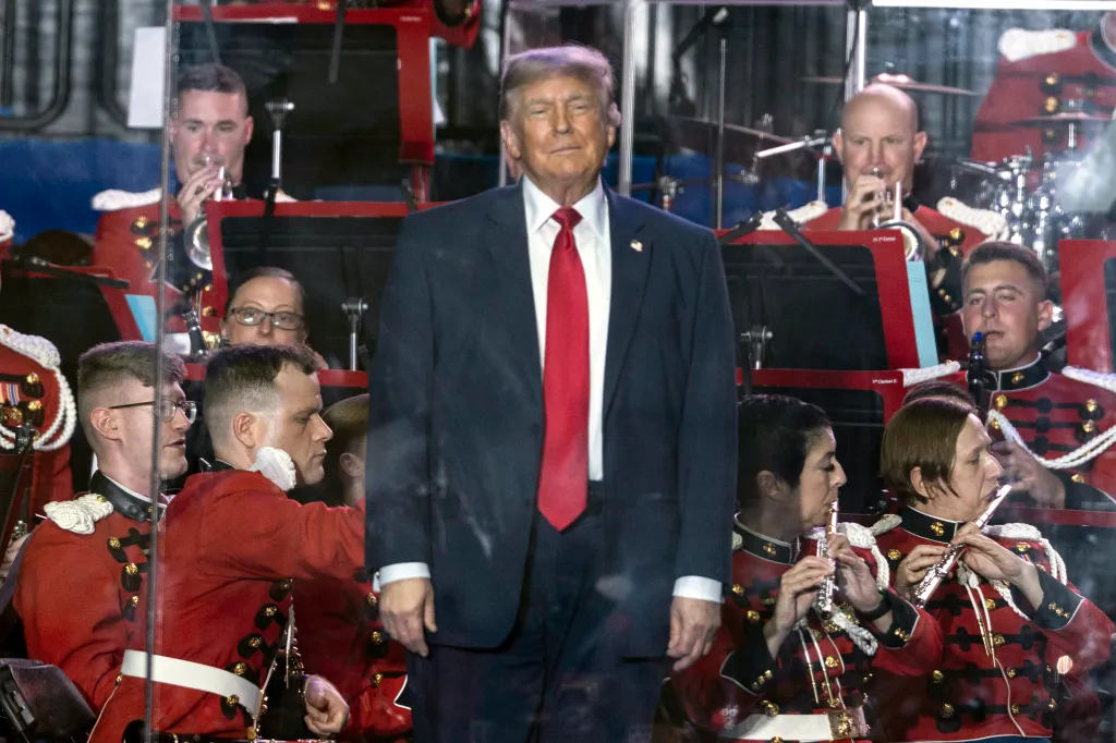 Donald Trump stands in front of a military band dressed in red and gold uniforms.