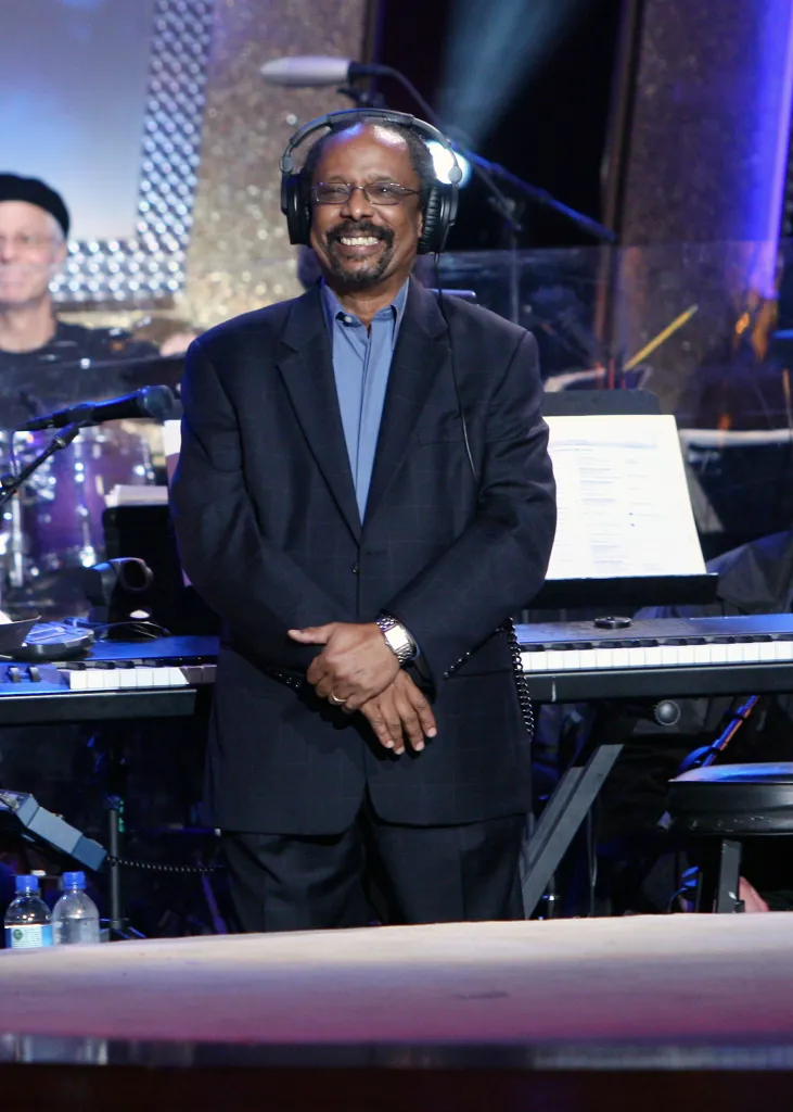 A man in a suit wearing headphones stands in front of a keyboard.