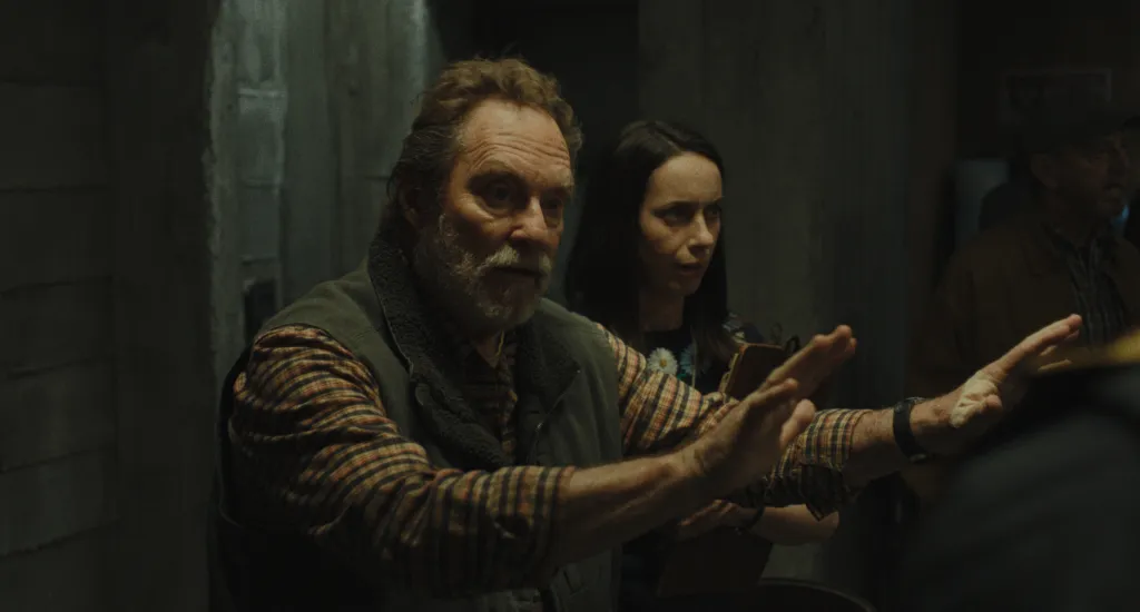 A man with a grey beard wearing a plaid shirt and vest speaks, hands outstretched, with a woman and another man behind him.
