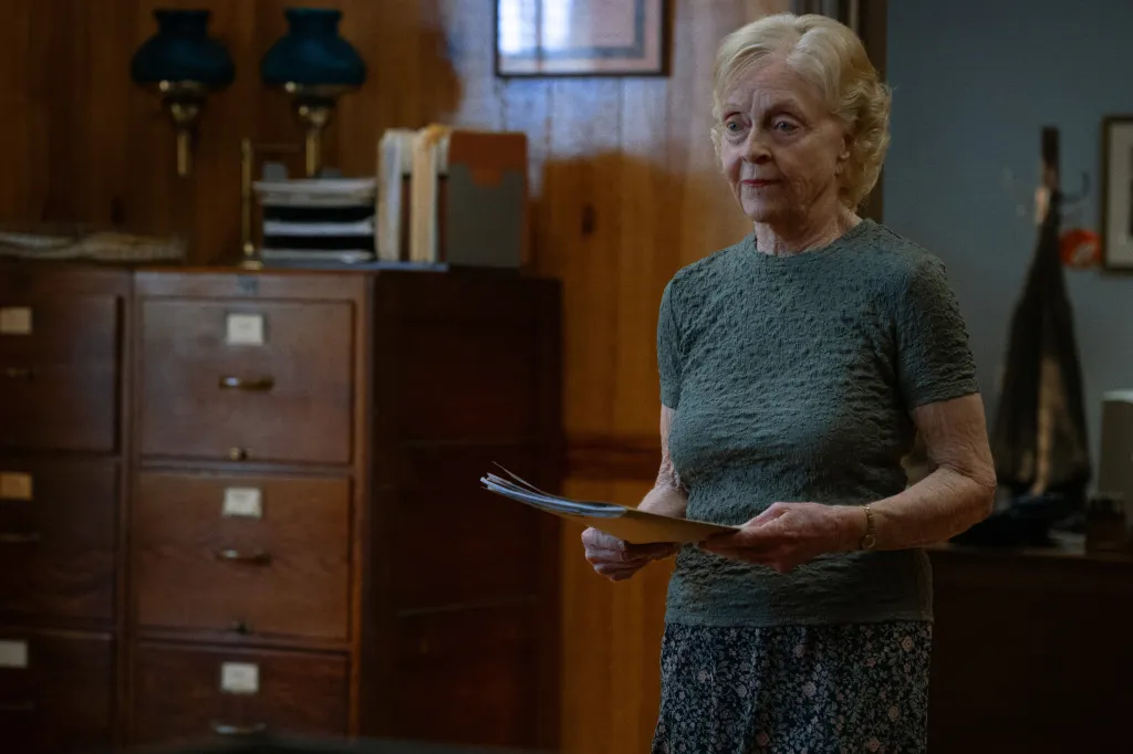 An older woman in a green top holds a folder and papers.