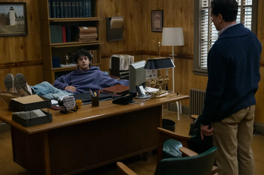 A young man with curly hair sits at a wooden desk with his feet up, talking to an older man whose back is to the camera.