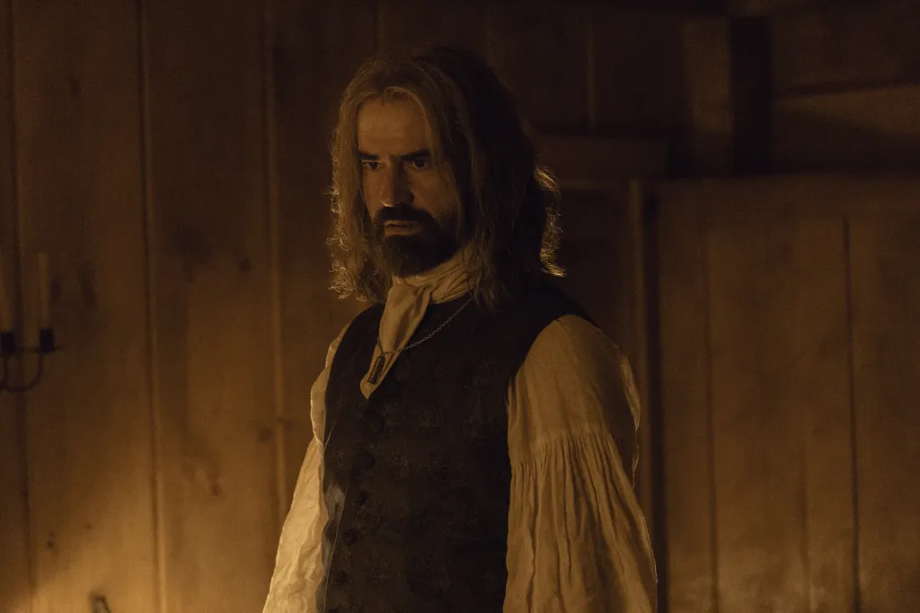 A man with long, light brown hair and a beard, wearing a period costume with a dark vest and white shirt, looks intently off to the left.