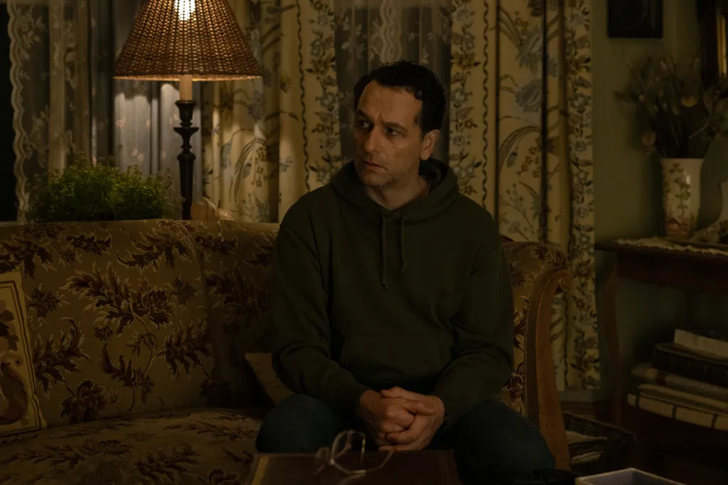 A man sits on a floral couch in a dimly lit room, looking away from the camera.