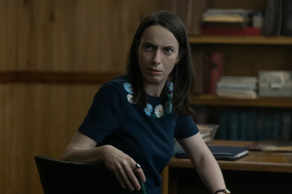 A woman with long dark hair and a daisy-patterned top looks intently to her left.