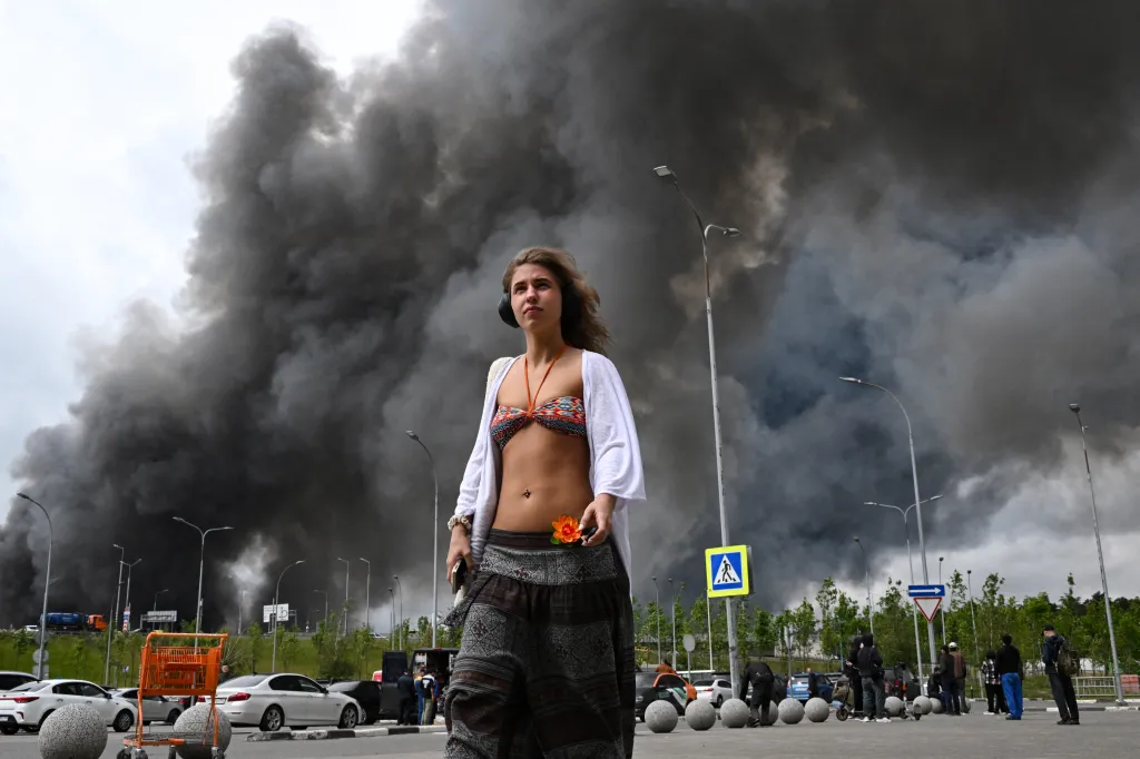 TOPSHOT - A woman walks outside a shopping mall as black smoke rises from the area of the Russian oil producer Gazprom Neft's Moscow oil refinery on the south-eastern outskirts of Moscow on June 18, 2026.