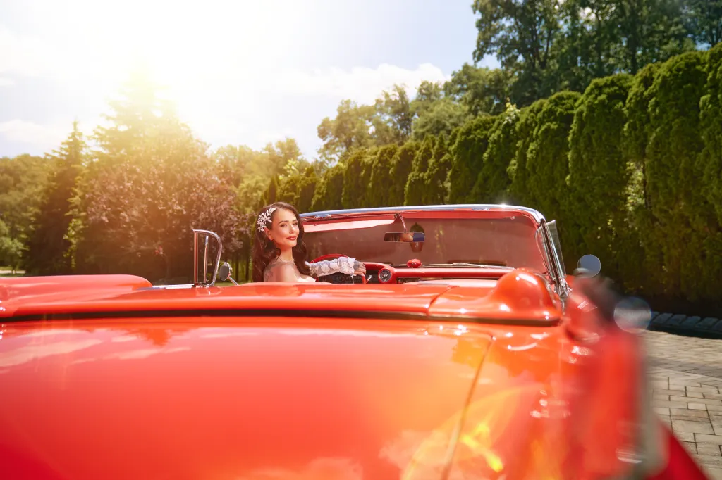 A woman with long wavy hair and a white floral headpiece smiles while sitting in a red convertible.