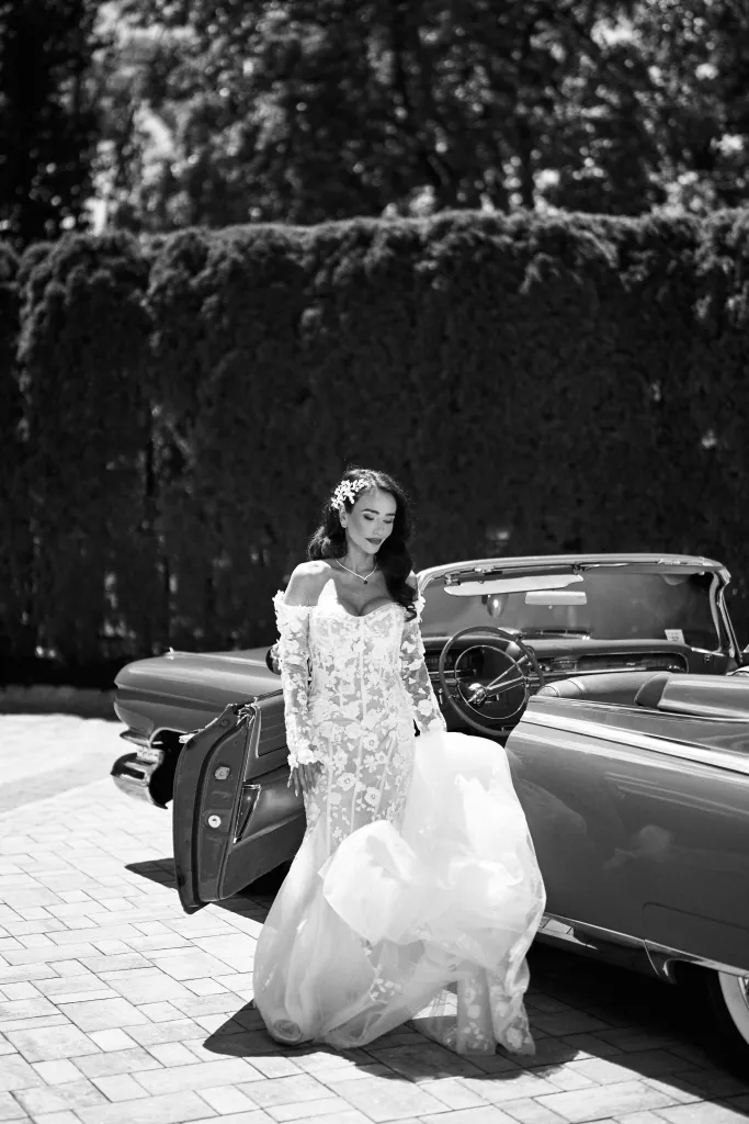 Black and white photo of a bride in an off-the-shoulder, long-sleeved, floral lace dress with a flowing train next to a classic convertible car.