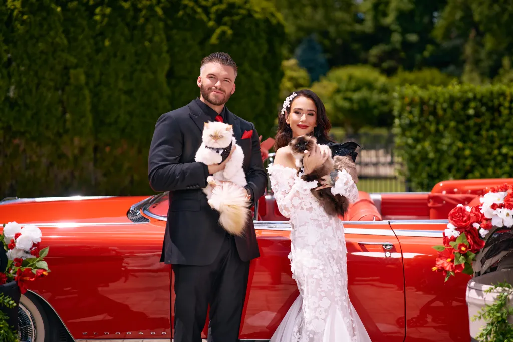 A man and woman, both holding cats, stand in front of a red convertible.