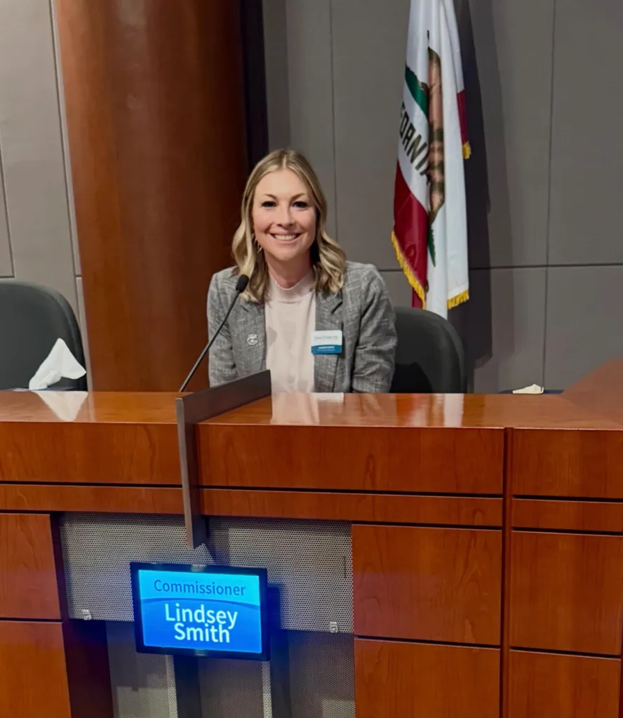 Lindsey Smith sitting at a large wooden desk with a microphone, in front of a California state flag.