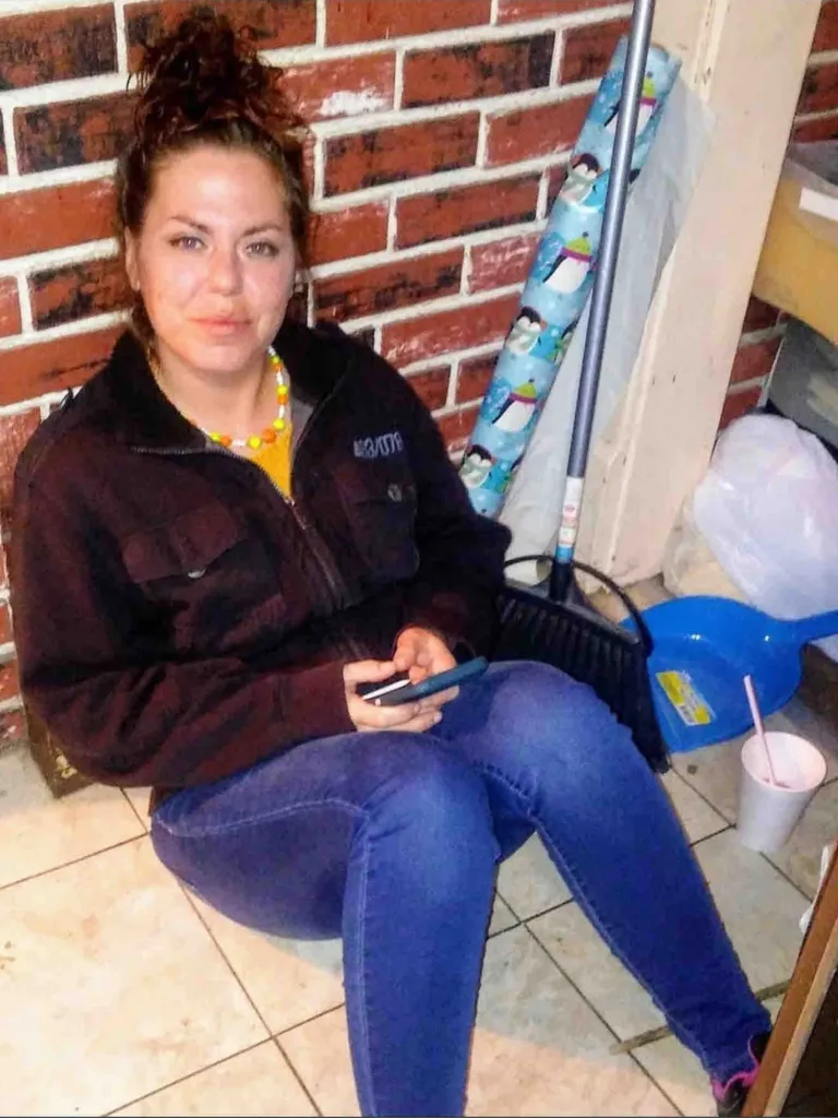 A woman with brown hair, dressed in a black jacket and jeans, sitting on a tiled floor with a brick wall behind her.