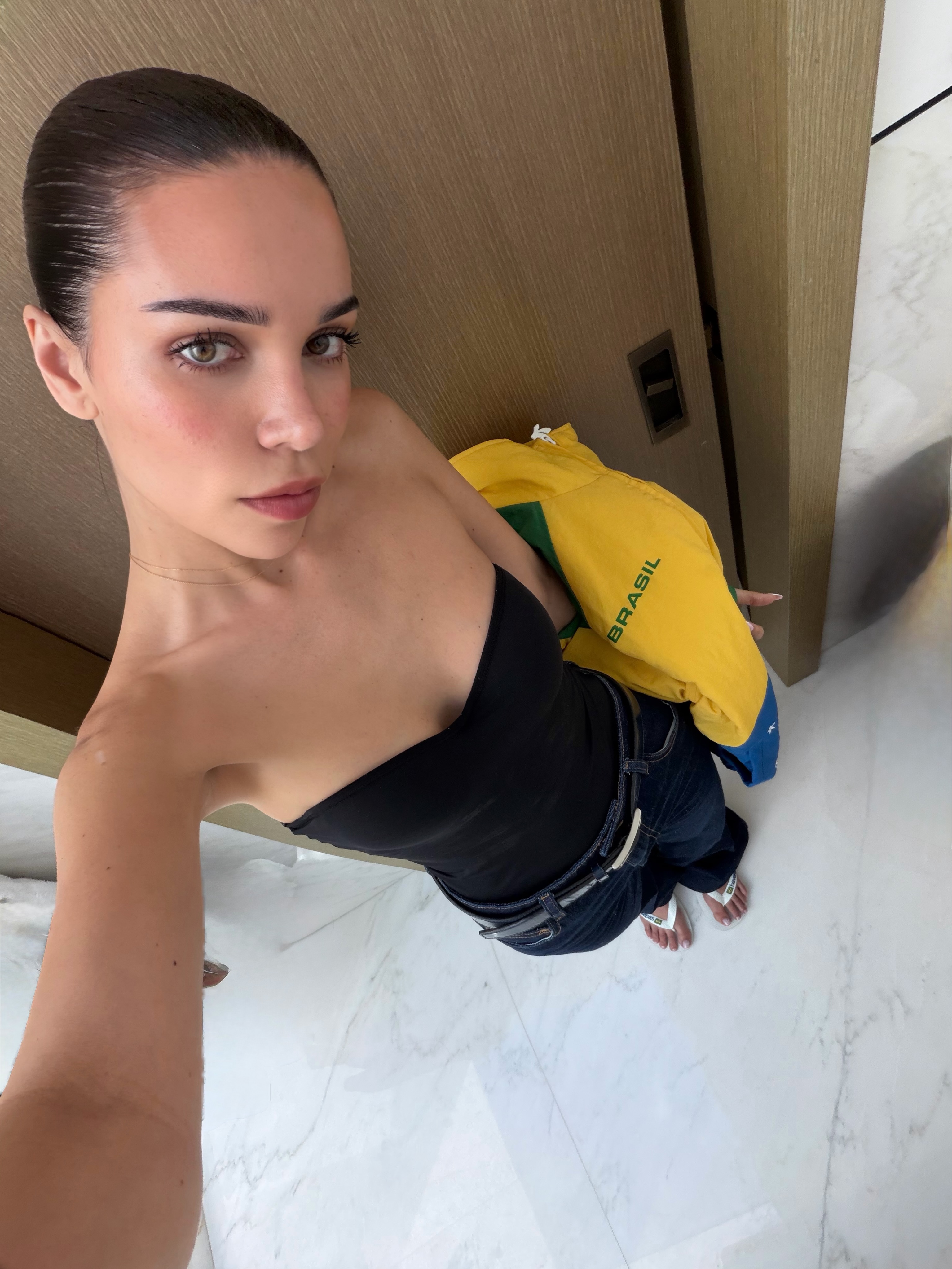 Selfie of Jackie Apostel wearing a strapless black top and dark jeans, holding a yellow jacket that says 
