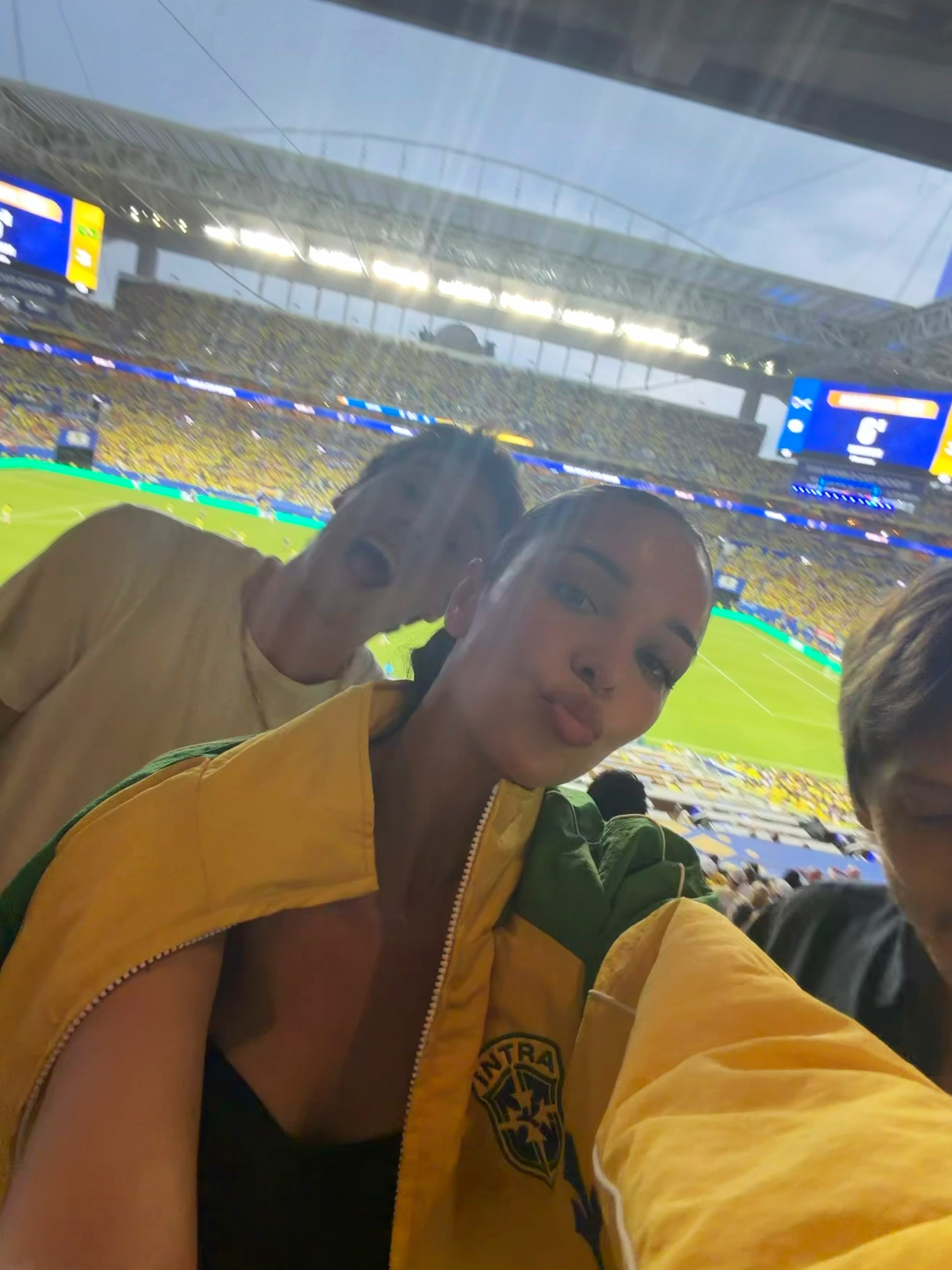 A couple takes a selfie at a soccer match, with the woman in a yellow and green jacket puckering her lips.
