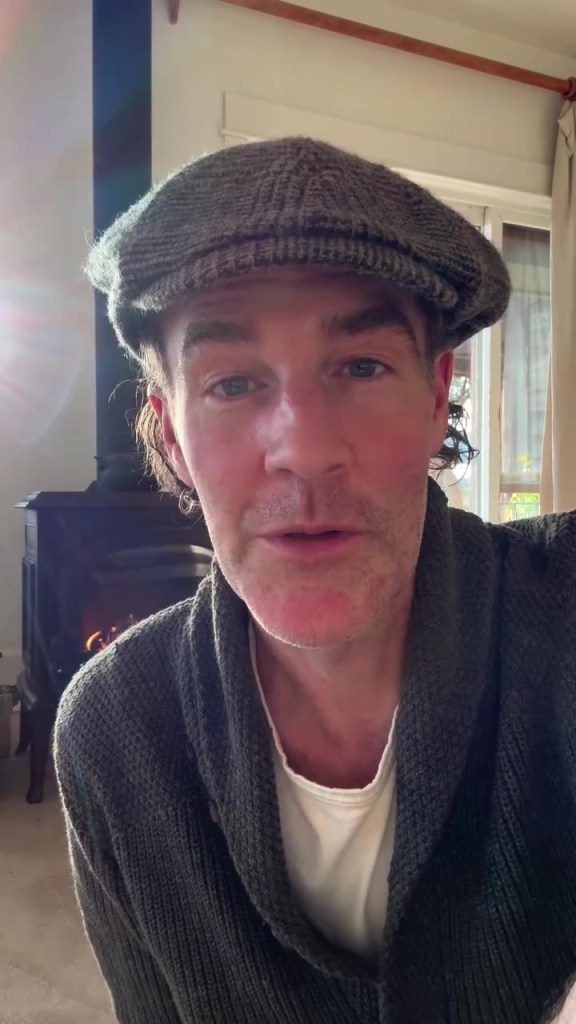 James Van Der Beek wearing a flat cap and a gray cardigan in front of a fireplace.