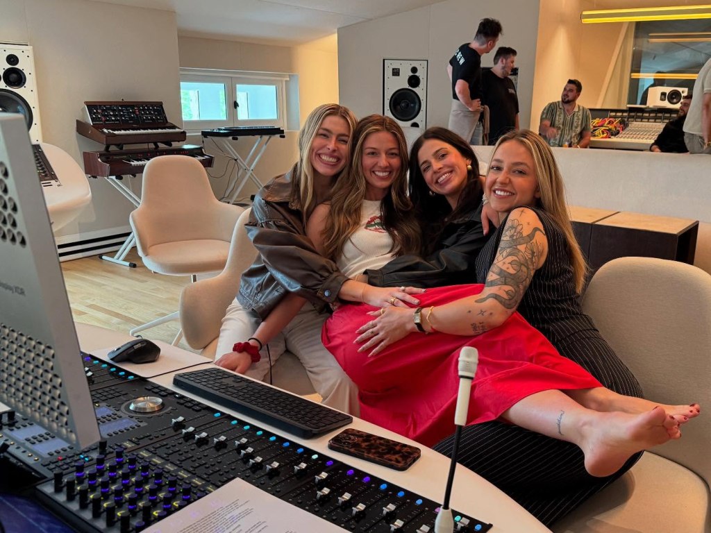 Four women smile in a recording studio, with audio equipment and other people in the background.
