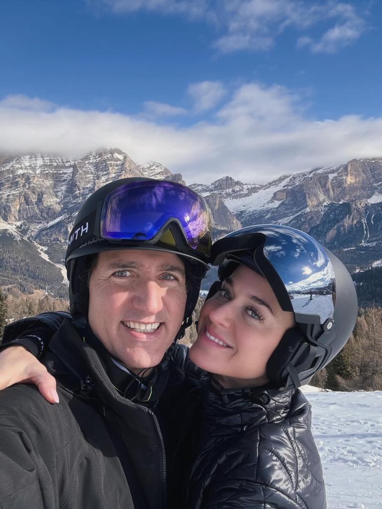 A smiling couple in ski helmets and goggles pose with snowy mountains in the background.