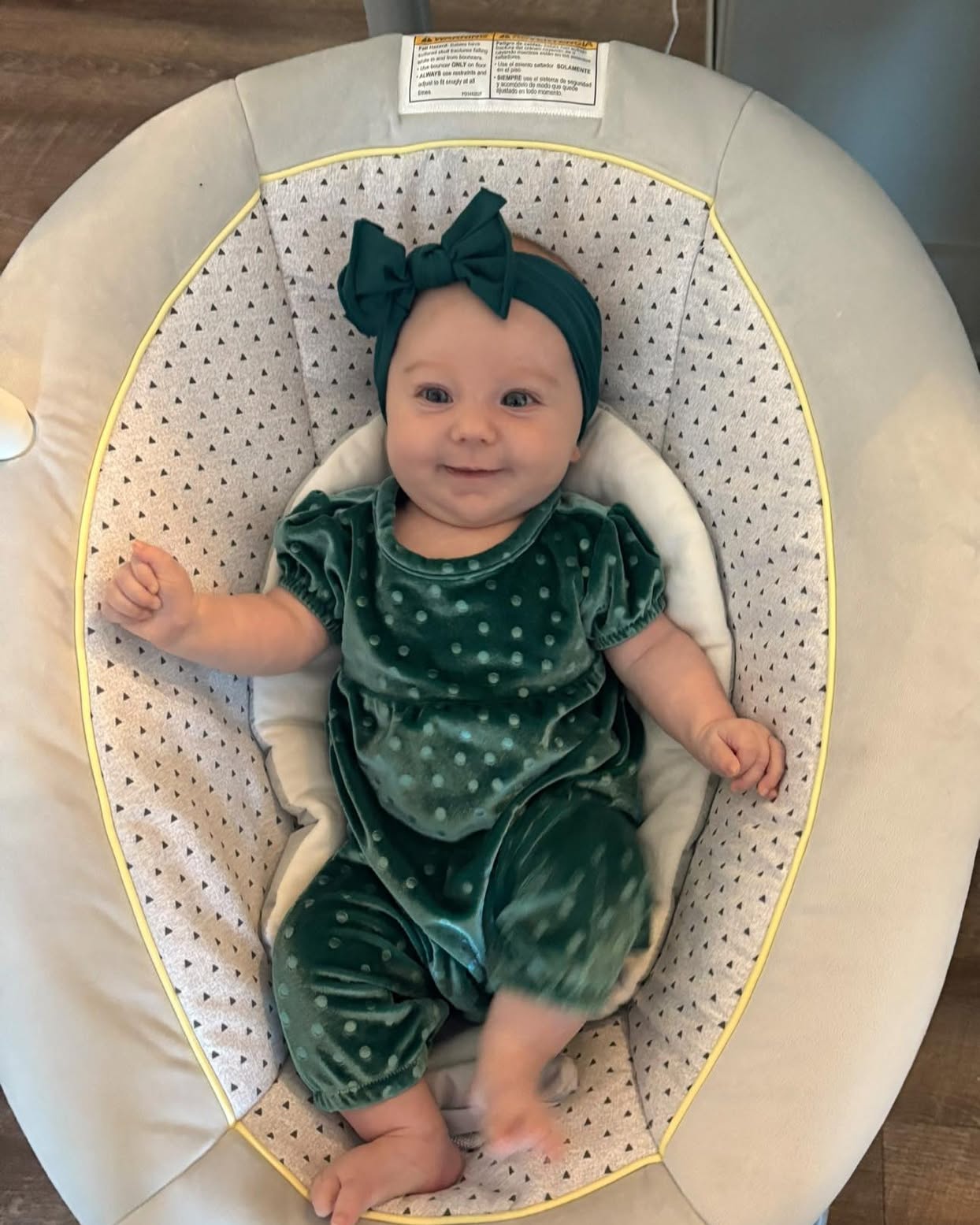 A smiling baby in a green polka-dotted romper and a matching bow headband sits in a bouncer.