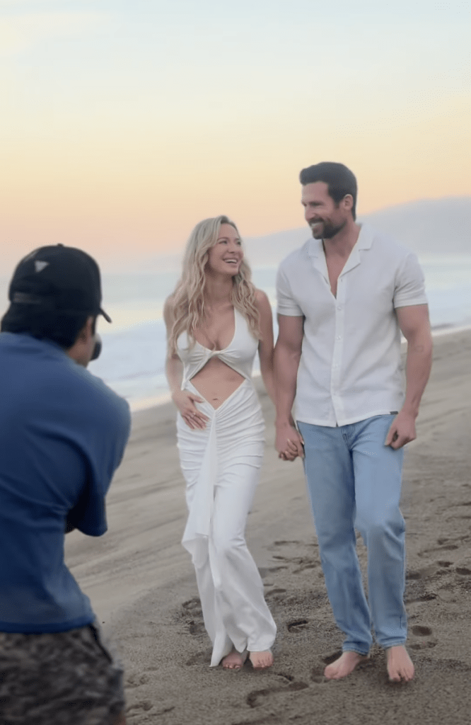 A woman in a white cutout dress and a man in a white shirt and blue jeans, holding hands and barefoot, walking on a beach while being photographed.