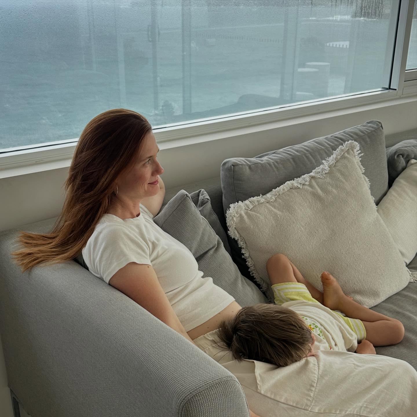 Bonnie Wright sits on a couch with a child sleeping on her lap.