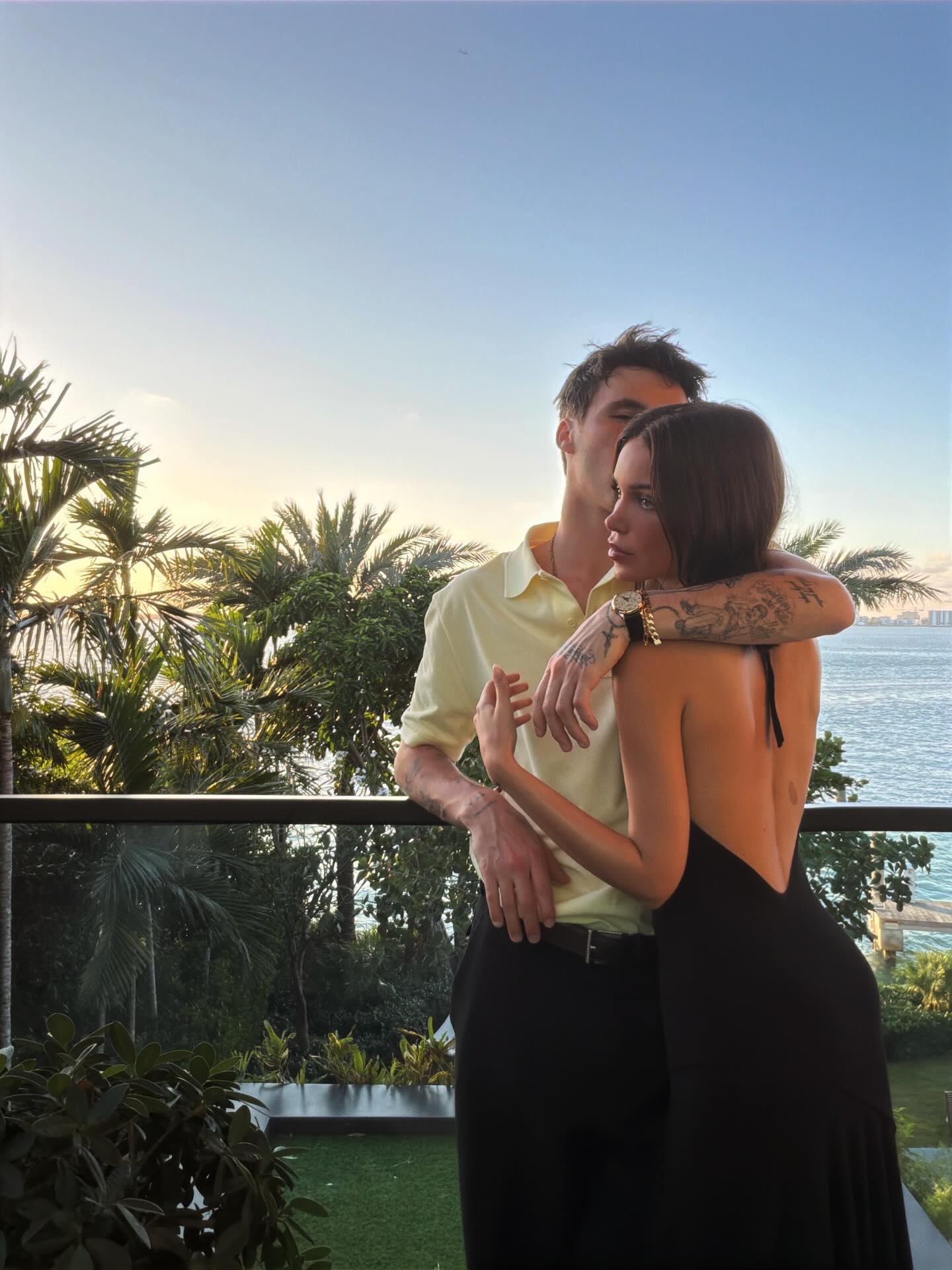 Cruz Beckham and Jackie Apostel embrace on a balcony with ocean and palm tree views at sunset.
