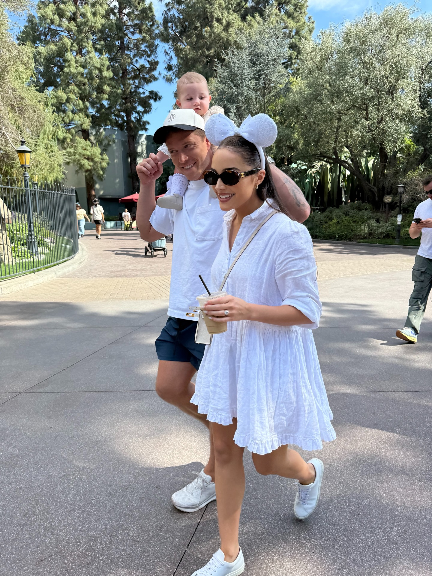 Olivia Culpo walking with a man carrying a baby on his shoulders, all three wearing white, in an outdoor park.