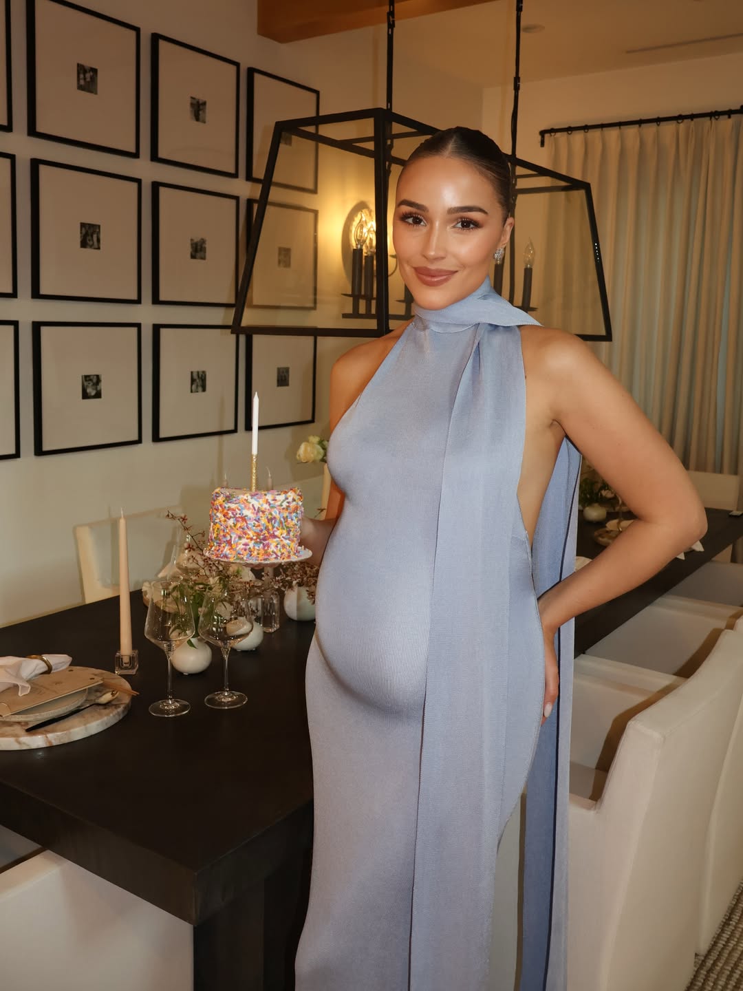Olivia Culpo in a light blue dress, celebrating her birthday with a small cake on a dining table.