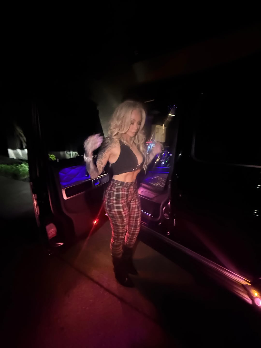 Bunnie XO in a low-cut top and plaid pants, standing next to a vehicle.
