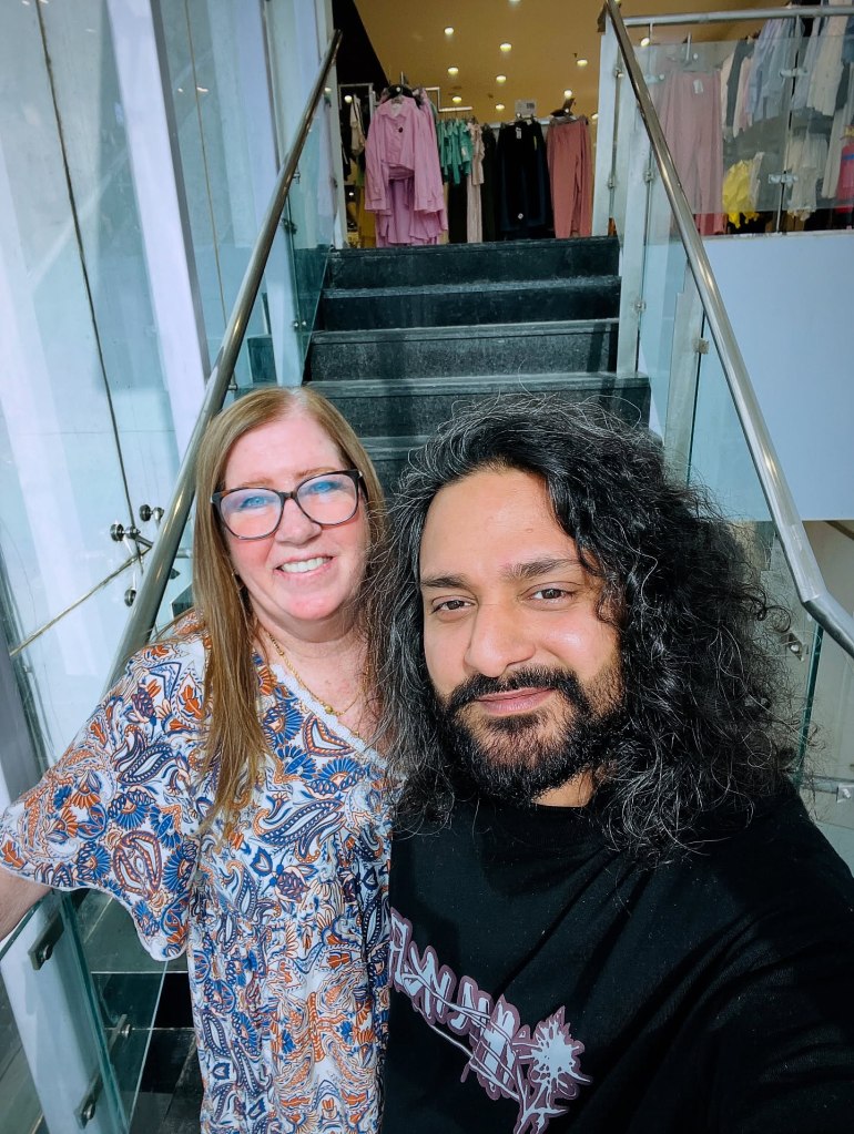 A man with long dark hair and a beard and a smiling woman with glasses pose in front of a stairway.