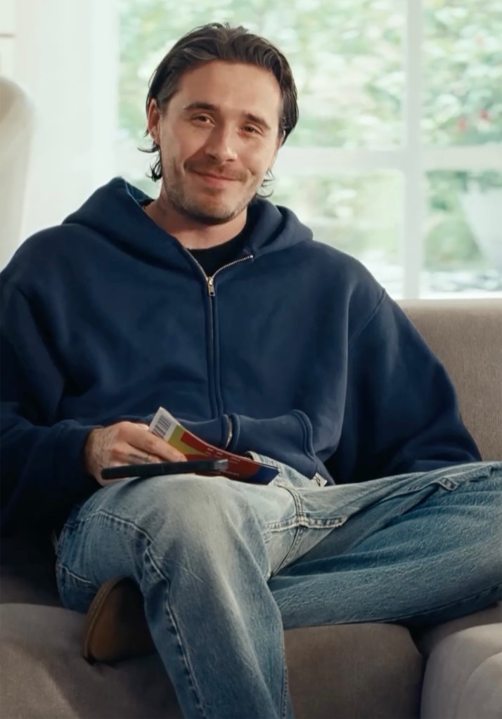 Brooklyn Beckham, in a blue hoody and blue jeans, smiling and holding a DoorDash menu.